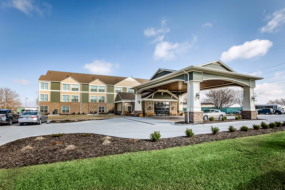 Exterior view of Remington Heights Retirement Community building with a covered entrance, parking area with cars, landscaped garden beds, and a clear blue sky.