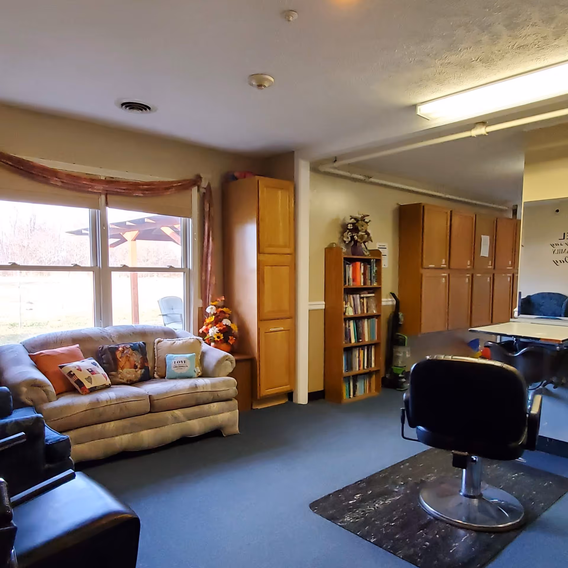 A cozy common area with a beige couch adorned with decorative pillows, a black chair, a bookshelf filled with books, wooden cabinets along the wall, and a window showing an outdoor pergola and chair. The room has blue carpet and neutral-colored walls.