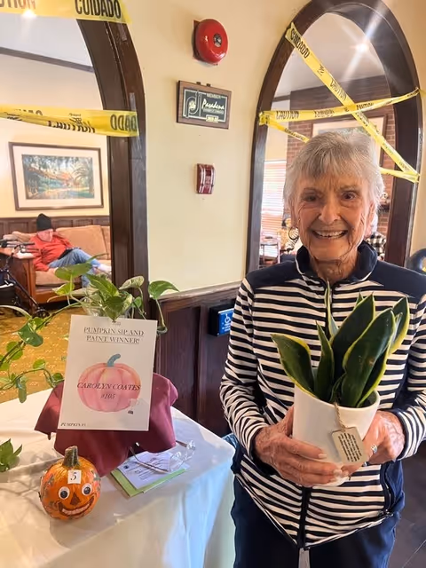 An elderly woman smiling and holding a potted plant indoors. Behind her, there is a table with a small decorated pumpkin and a sign that reads 'Pumpkin Sip and Paint Winner! Carolyn Coates #105'. The room has arched doorways with yellow caution tape across them. Another elderly person is sitting on a couch in the background.