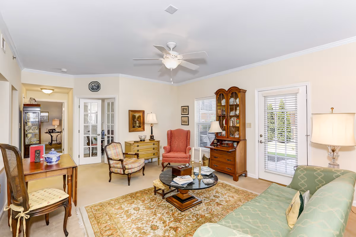 A bright and cozy living room in an active adult community featuring a green patterned sofa, a round coffee table with books and candles, a red armchair, a plaid armchair, wooden furniture including a china cabinet and a small dresser, a ceiling fan with light, and a door with window blinds leading outside.