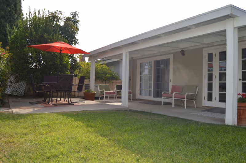 Outdoor patio area with a covered porch featuring chairs with cushions, a glass door entrance, and a separate seating area with a table and chairs under a red umbrella. The area is surrounded by greenery and a well-maintained lawn.