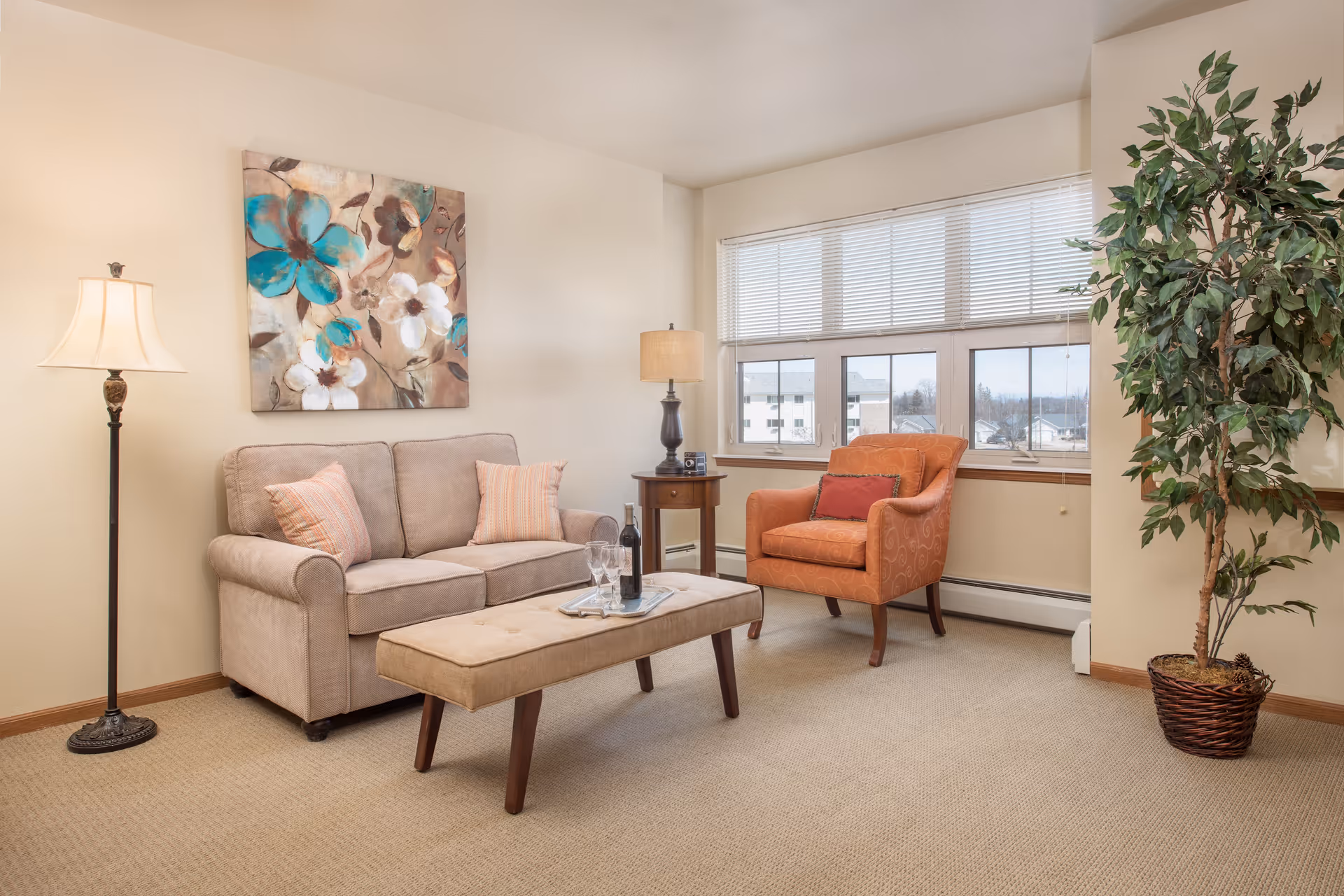 A cozy living room with a beige sofa adorned with two striped pillows, an orange armchair with a red cushion, a rectangular beige ottoman with a bottle of wine and two glasses on a tray, a tall floor lamp, a table lamp on a small wooden side table, a large window with blinds, a potted plant in the corner, and a floral painting on the wall.