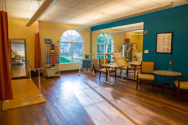 A bright physical therapy or rehabilitation room with wooden flooring, large arched windows letting in natural light, exercise equipment including parallel bars and weights, chairs, a small round table with a flower vase, and a large mirror on one wall.
