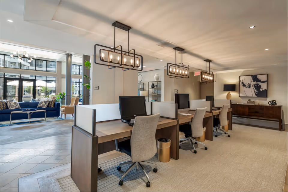 Modern office area with four workstations equipped with computers and beige swivel chairs. The space features contemporary pendant lighting, a carpeted floor, and a sideboard with a lamp and abstract artwork on the wall. In the background, there is a lounge area with blue sofas, beige armchairs, large windows, and natural light.
