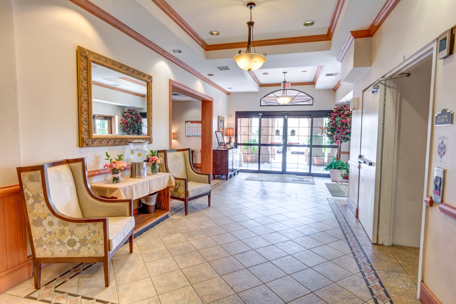 A bright and spacious lobby area with tiled floors, two upholstered chairs, a table with a water dispenser and flower vases, a large decorative mirror on the wall, potted plants, and glass double doors leading outside. The ceiling has recessed lighting and a hanging light fixture.