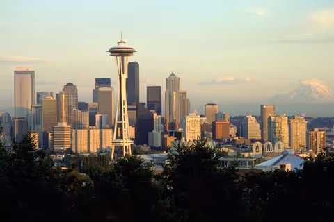 View of the Seattle skyline at sunset with the Space Needle prominently in the foreground and Mount Rainier visible in the background, framed by trees.