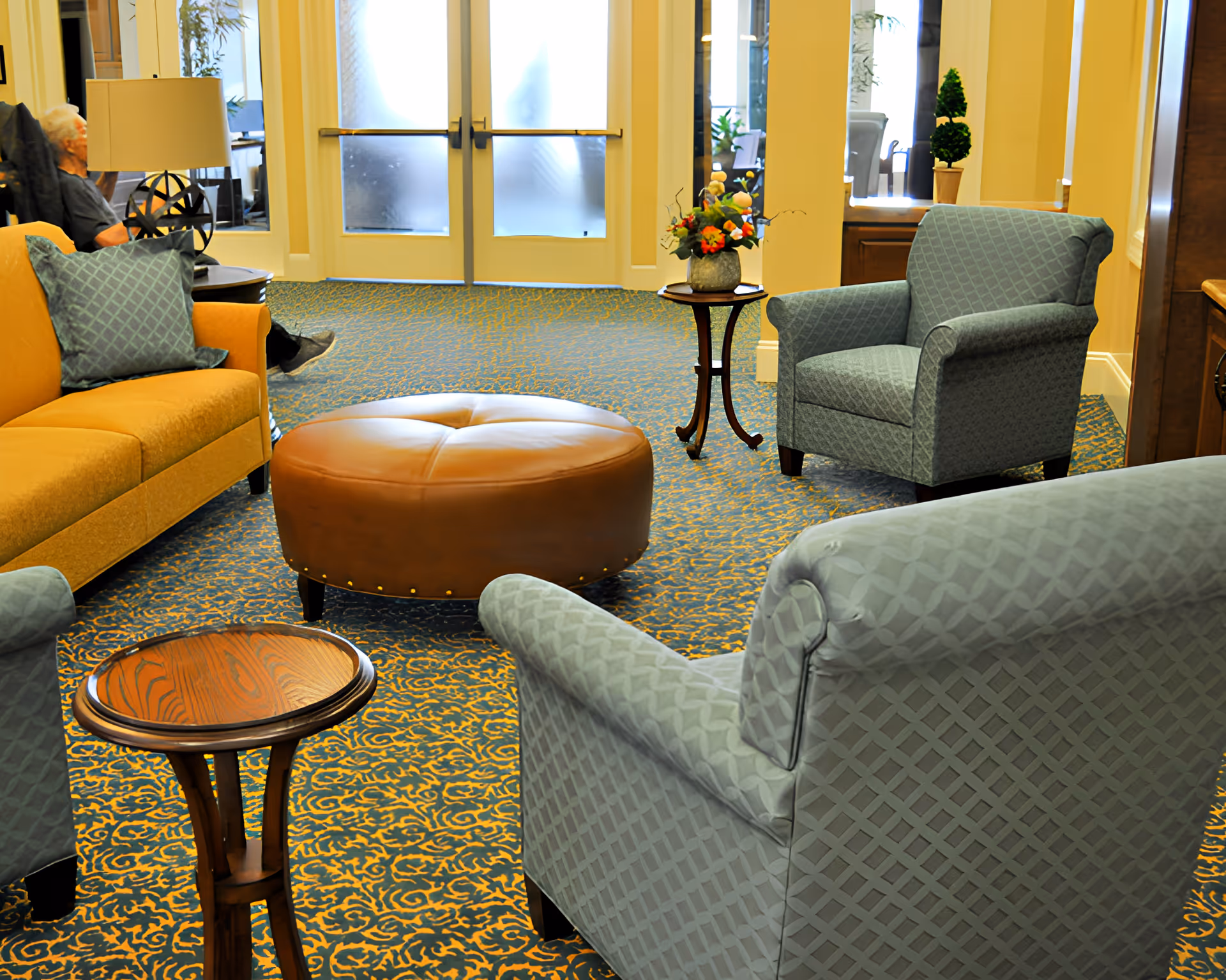 A cozy living room area in a senior living facility with patterned blue armchairs, a mustard yellow sofa with a blue cushion, a round brown leather ottoman, and two small wooden side tables. There is a floral arrangement on one side table and a lamp on another. In the background, a senior person is seated near a window with frosted glass doors.