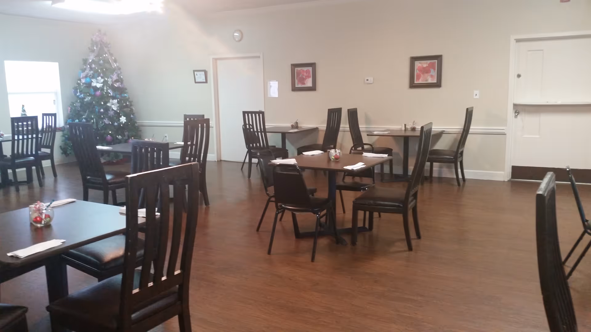A dining room with several dark wooden tables and chairs arranged neatly. Each table has a small centerpiece and napkins. There is a decorated Christmas tree in the corner near a window, and two framed floral pictures hang on the beige walls.