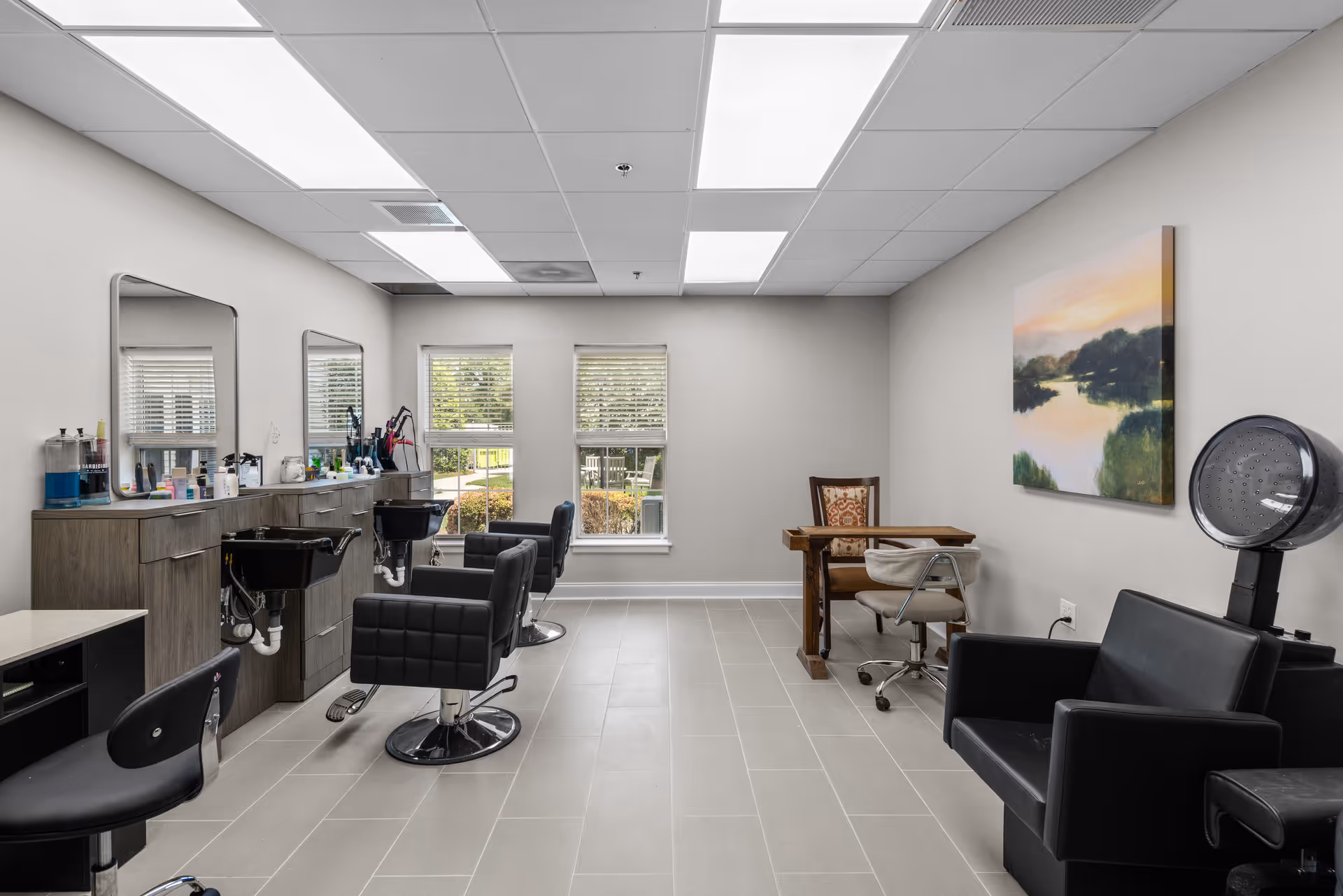 Interior view of a salon area in a senior living facility with black salon chairs, hair washing sinks, a hair dryer chair, a small wooden table with a chair, and a painting of a landscape on the wall. Large windows allow natural light to enter the room.