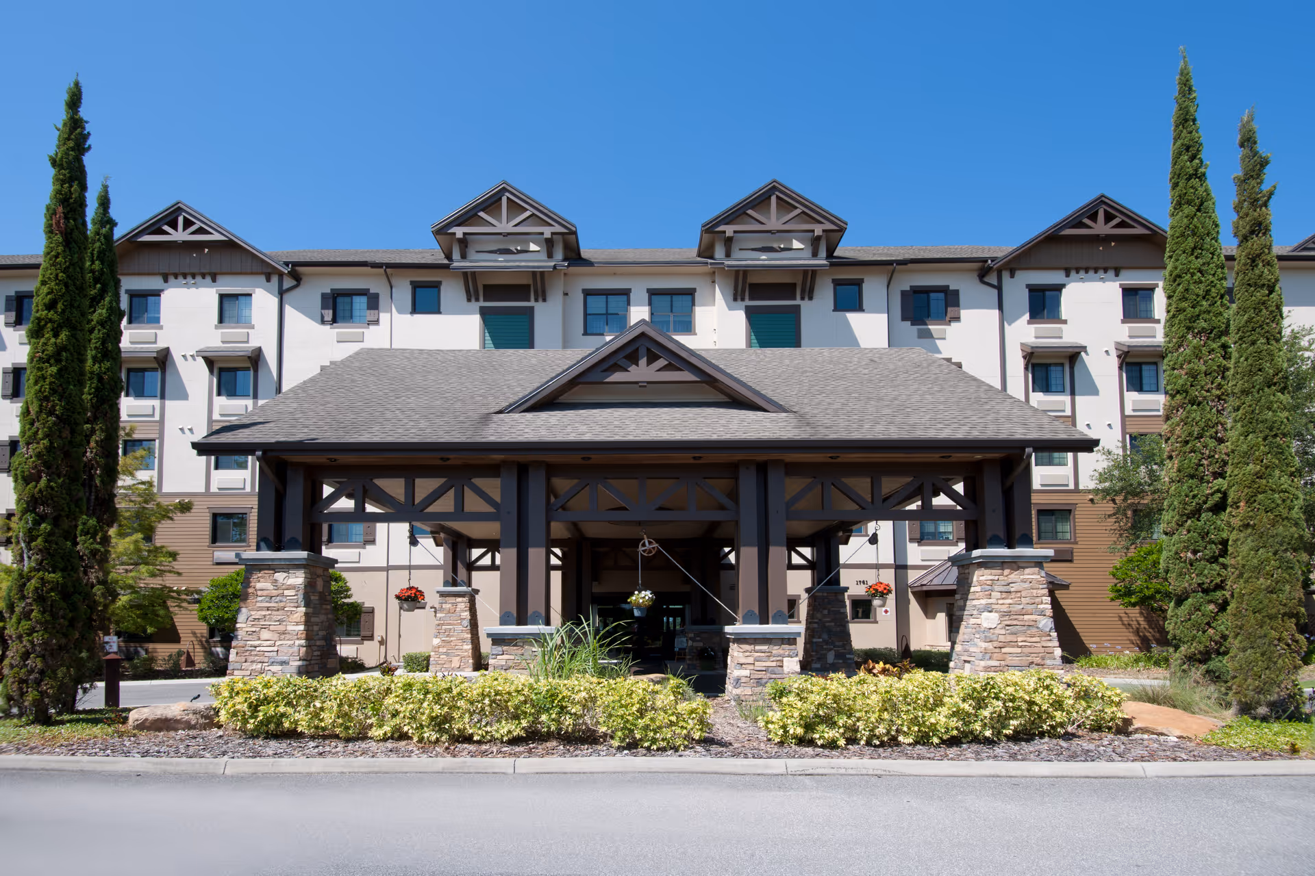 Front exterior view of a multi-story senior living facility with a large covered entrance supported by stone pillars, surrounded by neatly trimmed bushes and tall trees under a clear blue sky.