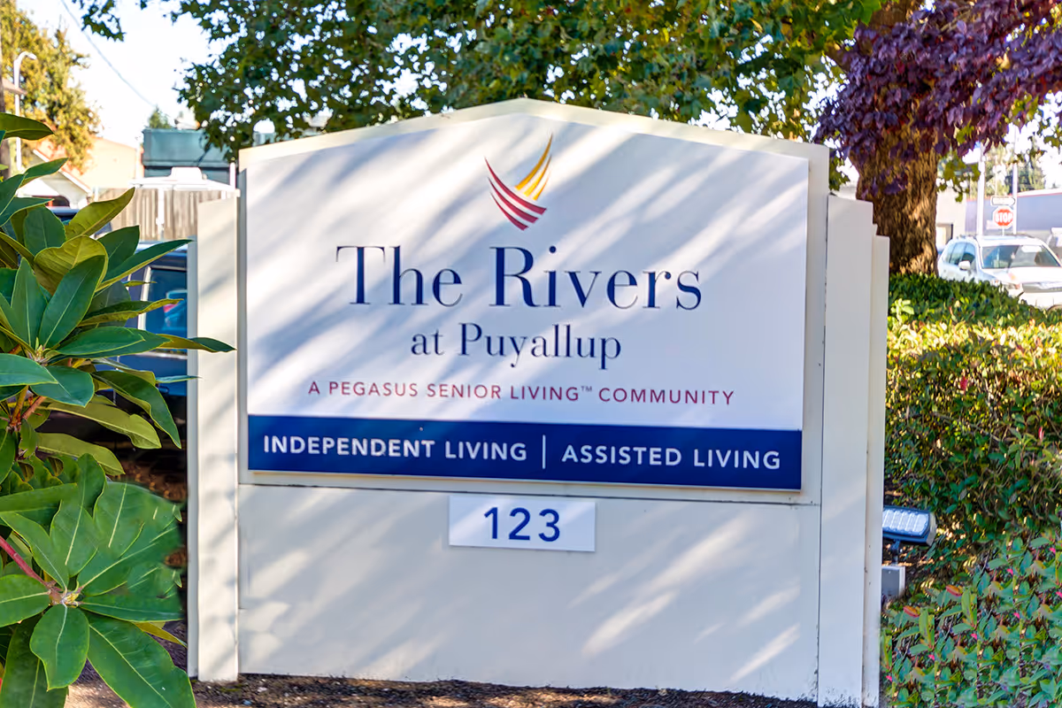 Outdoor sign for The Rivers at Puyallup, a Pegasus Senior Living community offering independent living and assisted living, surrounded by greenery and trees.