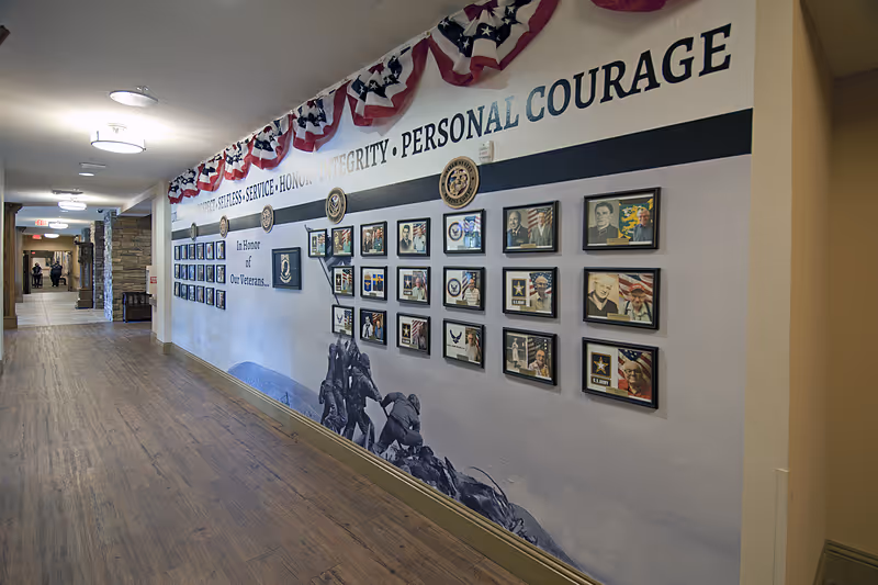 A hallway in a senior living facility with a wooden floor and stone pillars along the left side. The right wall features a large mural honoring veterans, decorated with patriotic red, white, and blue bunting. The mural includes framed photos, military insignias, and the words 'SELFLESS SERVICE • HONOR • INTEGRITY • PERSONAL COURAGE' along with 'In Honor of Our Veterans.'