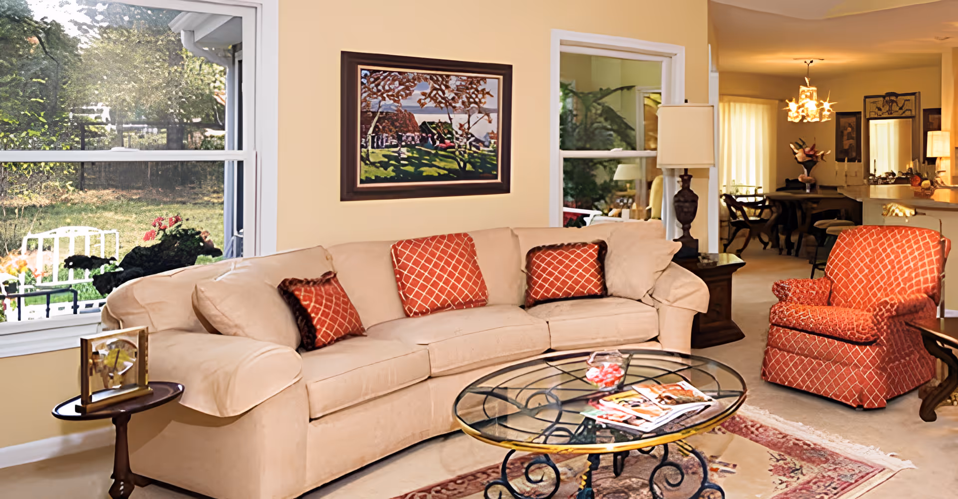 A cozy living room with a beige curved sofa adorned with red patterned cushions, a matching red armchair, a glass coffee table with magazines and a decorative bowl, a side table with a framed photo, and a painting on the wall. Large windows show a green outdoor garden area, and the room opens into a dining area with a chandelier and wooden furniture.