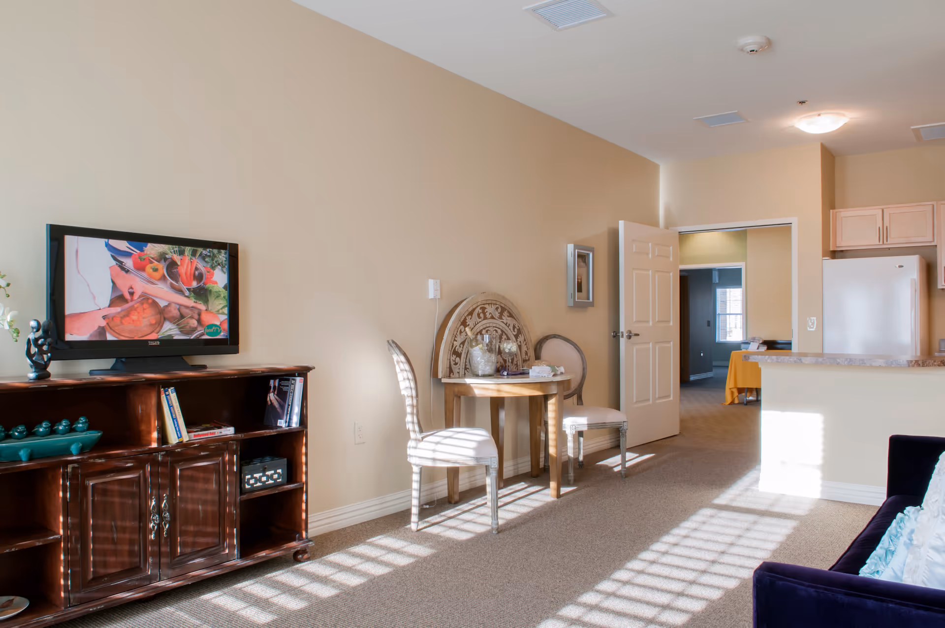 A bright living area with beige walls and carpeted floor. There is a wooden TV stand with a flat-screen TV showing a cooking program. Next to it is a small round wooden table with two upholstered chairs. An open door leads to another room, and part of a kitchen with light wood cabinets and a white refrigerator is visible. Sunlight streams through a window, casting shadows on the floor and furniture.