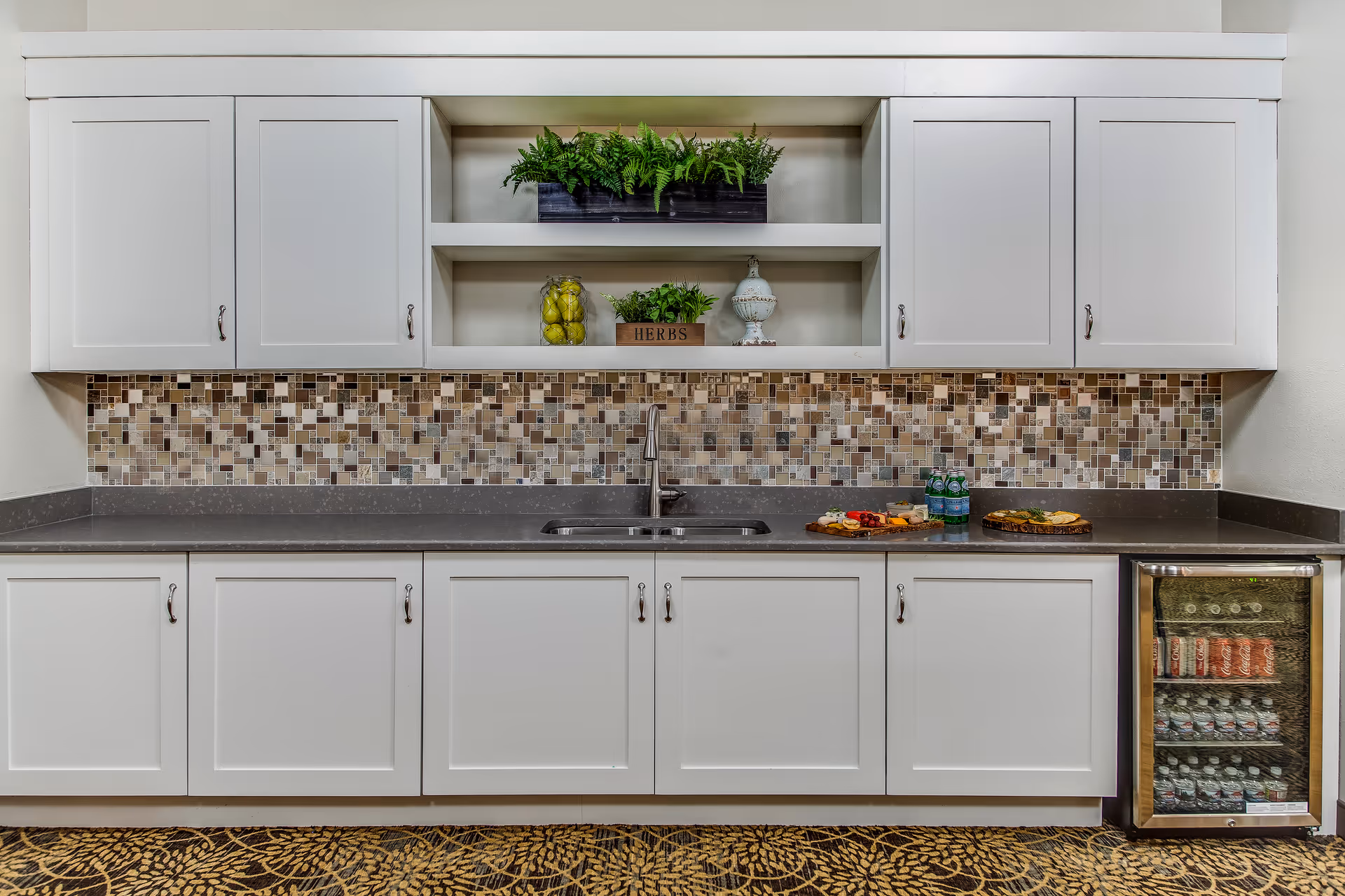 A modern kitchen area with white cabinets, a mosaic tile backsplash, a stainless steel sink with a faucet, and a small glass-front refrigerator stocked with beverages. The countertop has two wooden serving boards with snacks and bottled water. There are decorative plants and a jar of lemons on the open shelves above the sink.