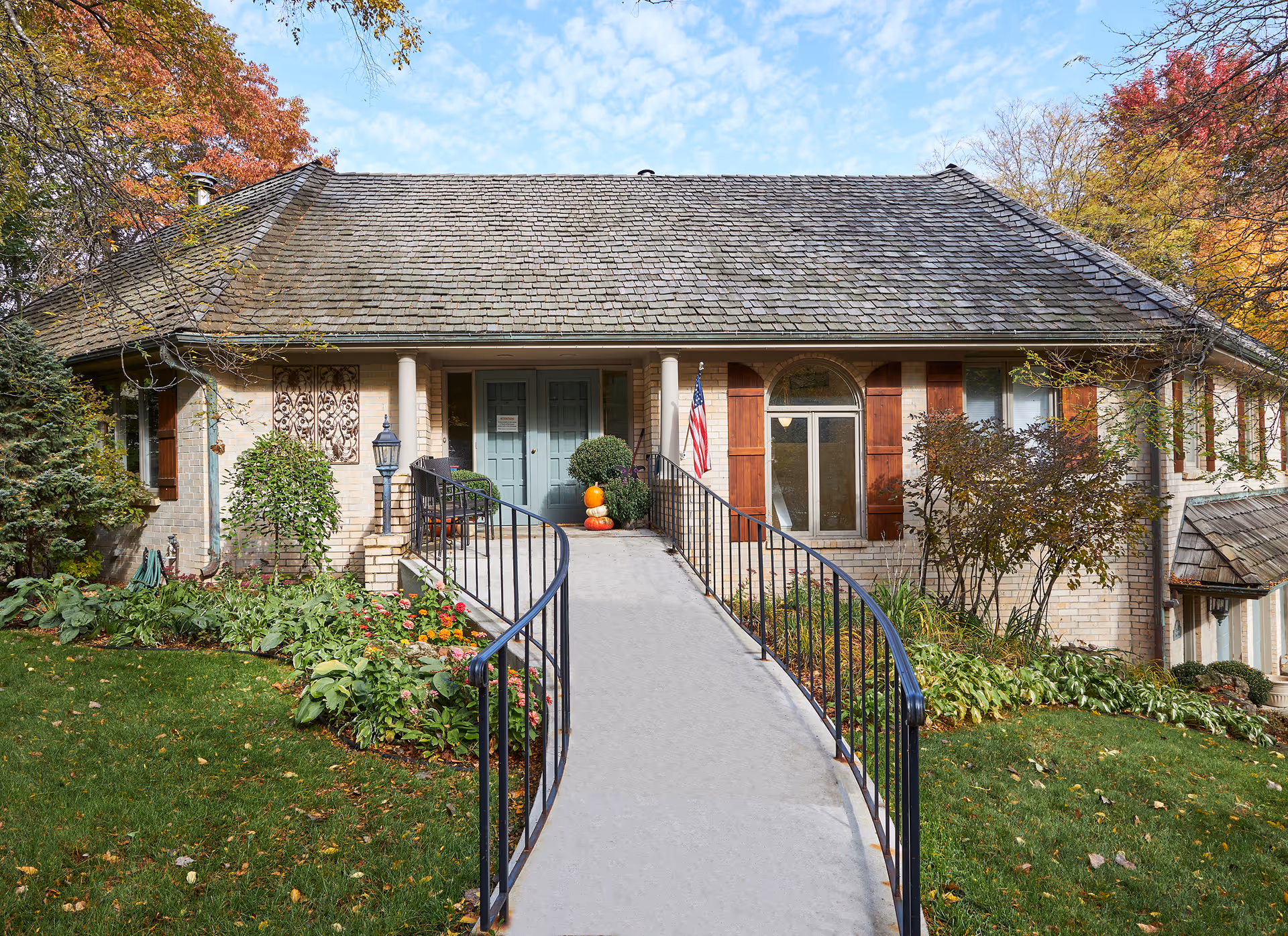 Front entrance of a single-story brick house with a ramp, iron railings, American flag and autumn landscaping.