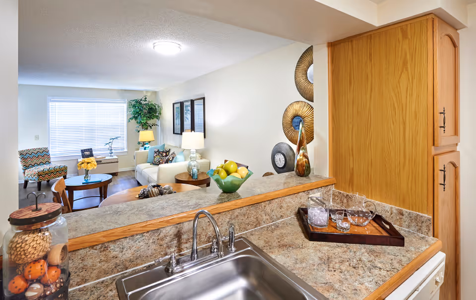 View from a kitchen sink area looking into a living room with a white sofa, a patterned armchair, a coffee table with a vase of yellow flowers, a side table with a lamp, and a large window with blinds. The kitchen counter has a bowl of fruit and a tray with glassware. Decorative wall art is visible on the right wall.