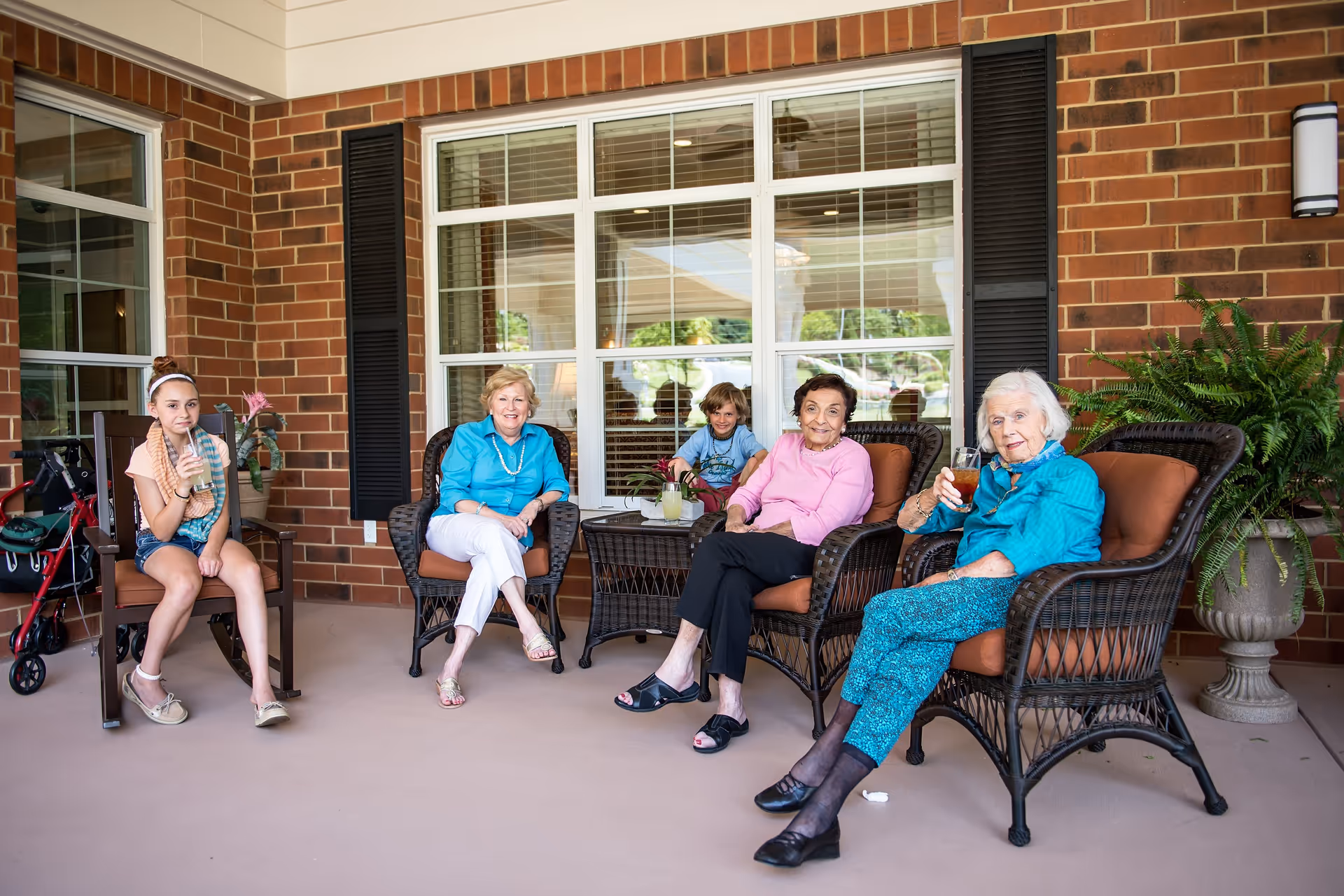 A group of elderly residents and two children seated on wicker chairs on a covered brick patio in front of a large window, some holding drinks.