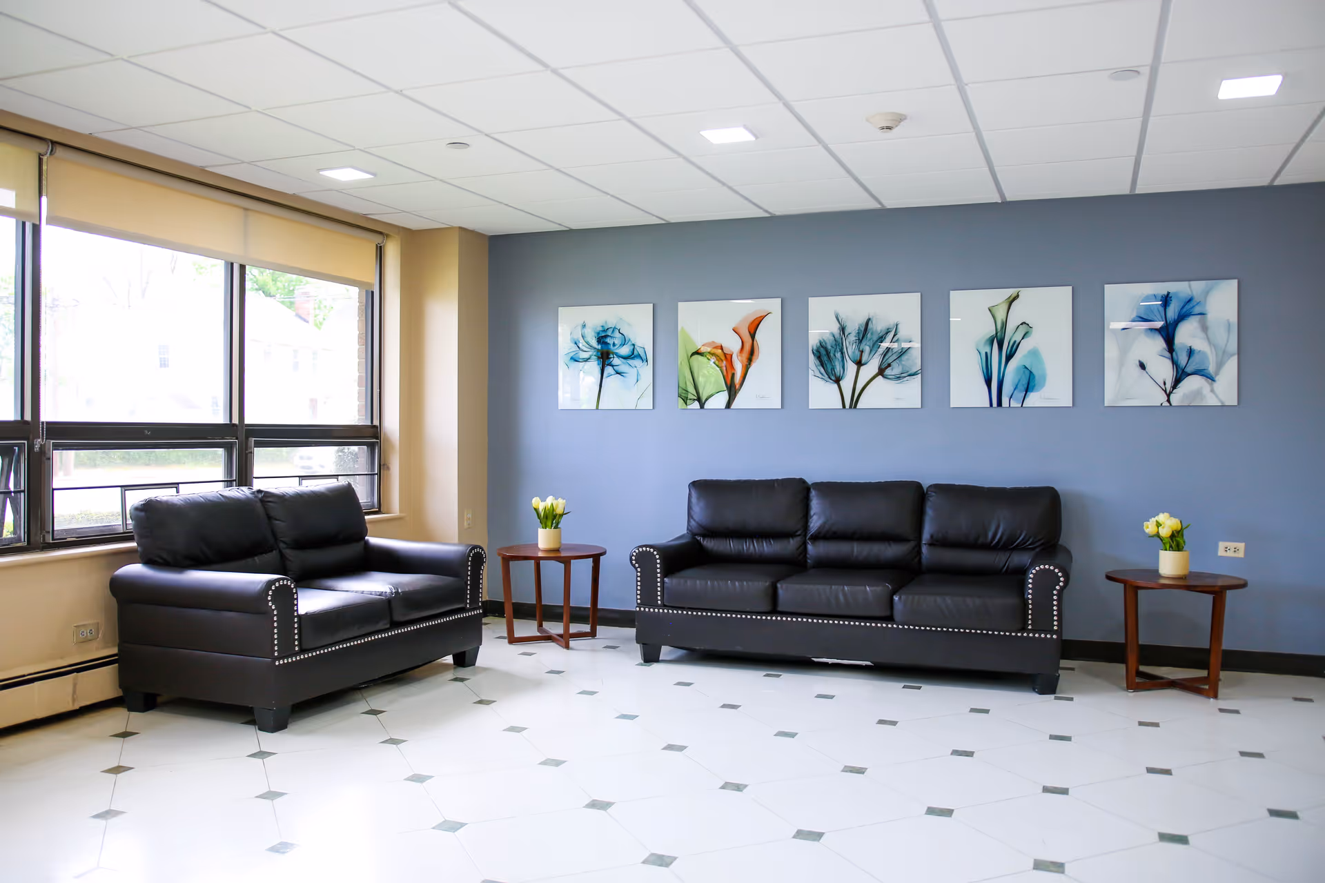 A bright and clean living room area with two black leather sofas, one two-seater and one three-seater, placed against a blue accent wall. Above the sofas are five framed floral artworks. There are two small wooden side tables with potted yellow flowers on each side of the sofas. Large windows with beige roller blinds allow natural light to fill the room. The floor is white with a subtle geometric pattern.