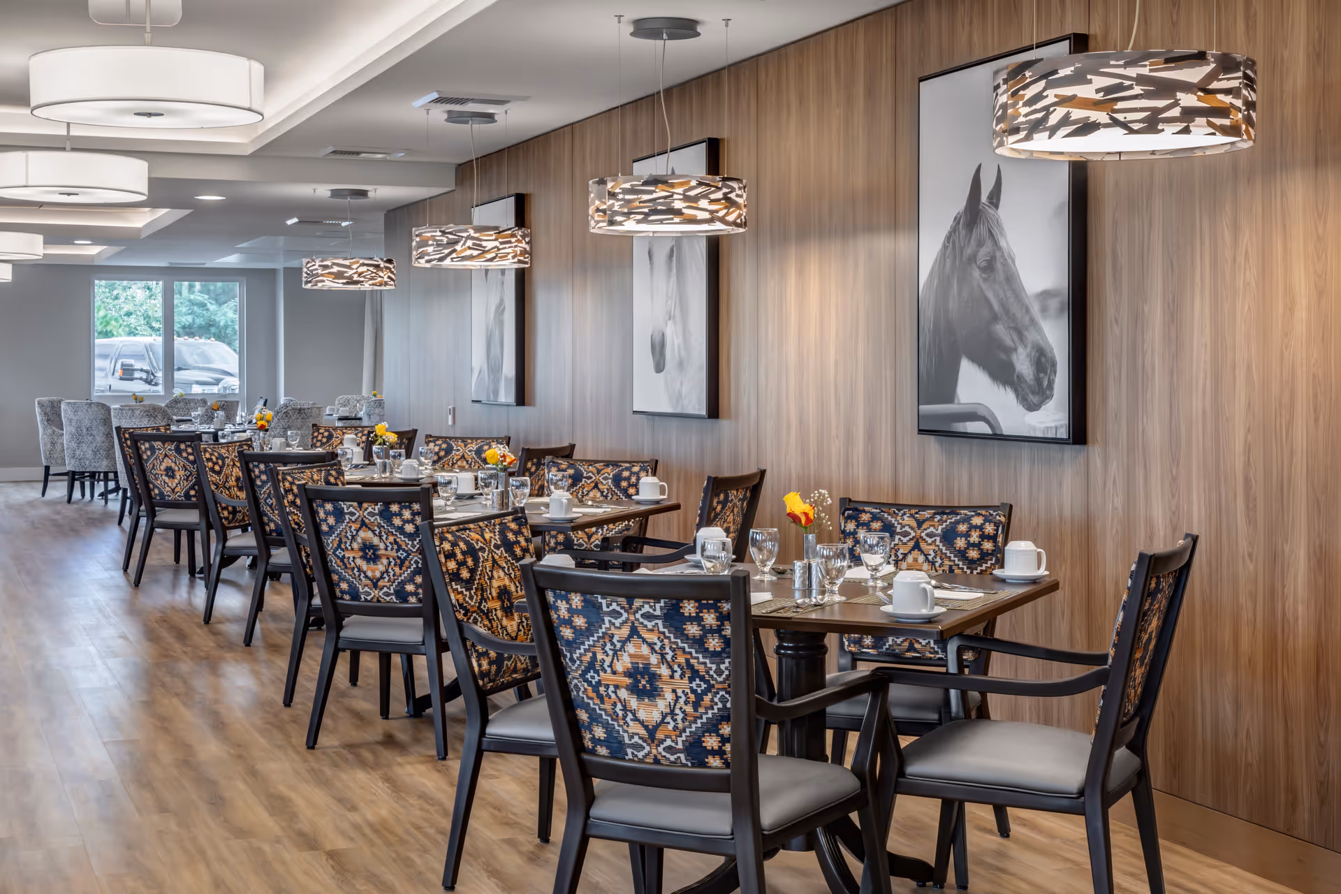 A modern dining room with multiple set tables and patterned chairs, pendant lights, and horse portraits on wood-paneled walls.