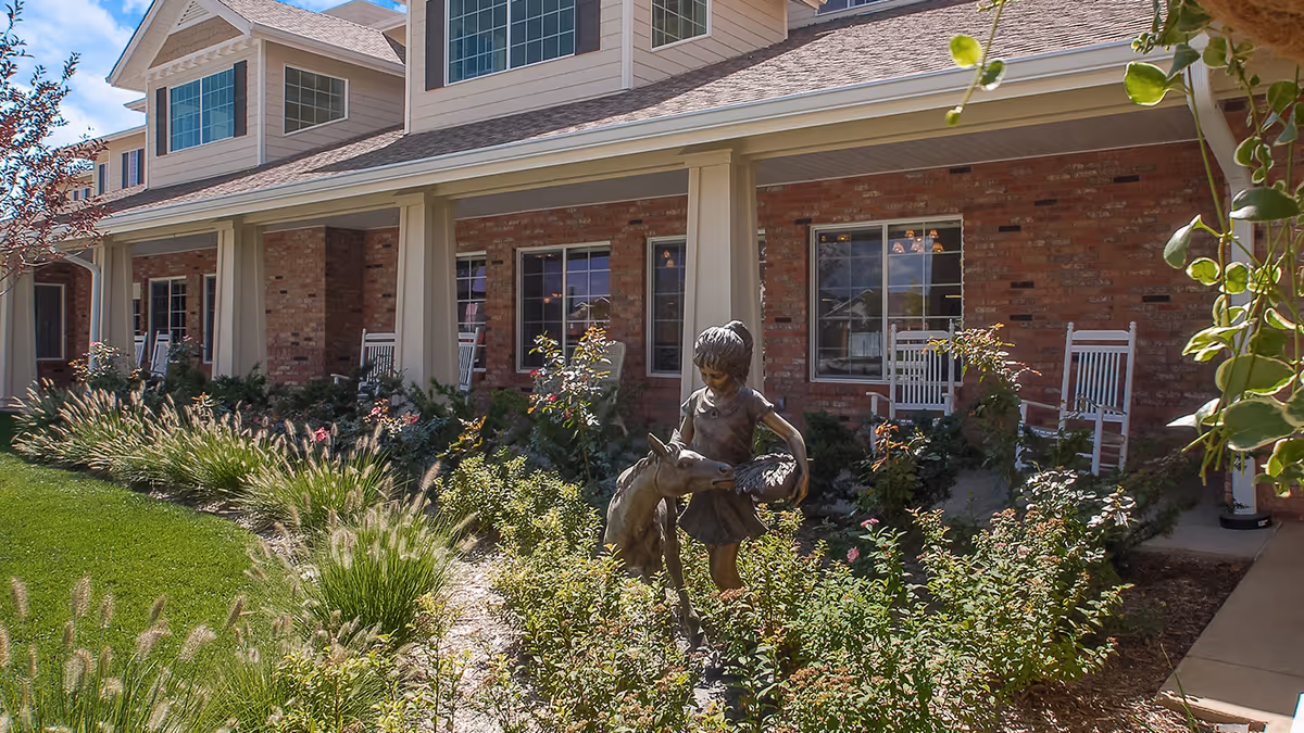 Exterior view of a senior living facility with a brick facade and beige siding. The building has a covered porch with white rocking chairs. In the foreground, there is a garden with various plants and a bronze statue of a young girl feeding a small horse or donkey.