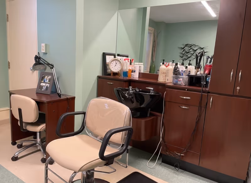 Small salon area with beige styling chairs, a black shampoo basin, wooden cabinetry and hair-care products on the counter.