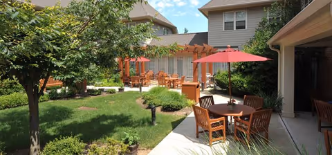 Outdoor patio area at a senior living facility with wooden tables and chairs, some shaded by red umbrellas, surrounded by green grass, trees, and shrubs, with a building in the background under a blue sky with scattered clouds.