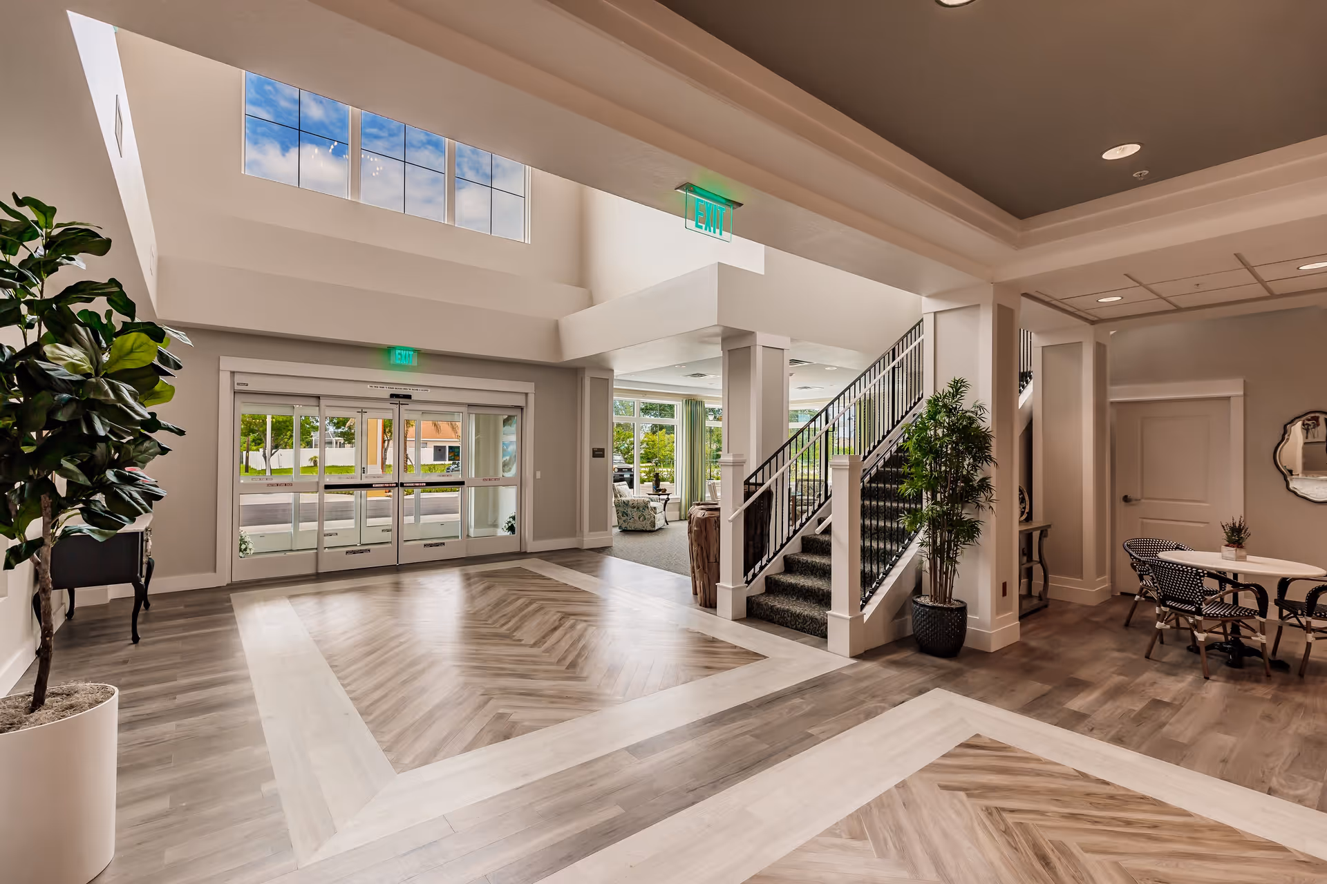 Spacious and well-lit interior lobby area of a senior living facility with large windows, a glass entrance door, a staircase with black railings, potted plants, and a small seating area with a round table and chairs.