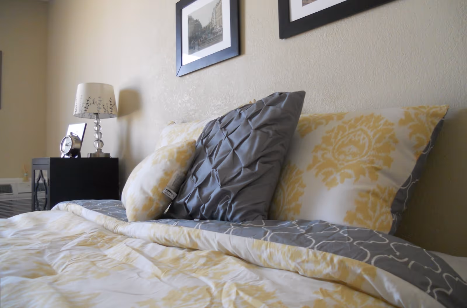 Close-up of a neatly made bed with decorative yellow and gray pillows and a bedside table with a lamp and alarm clock.