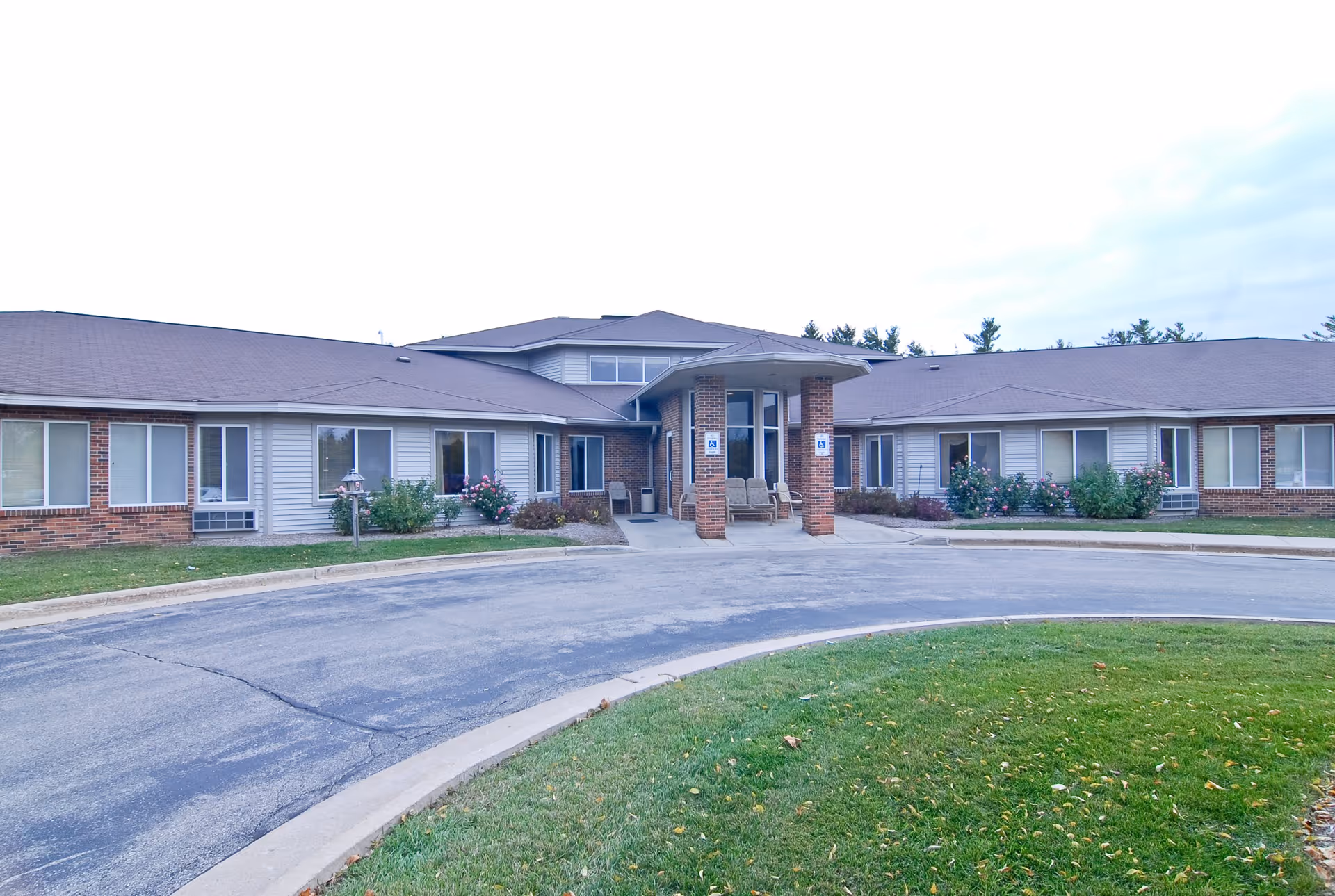 Front exterior view of a single-story senior living facility building with a covered entrance supported by brick columns, surrounded by a circular driveway and landscaped with grass and bushes.