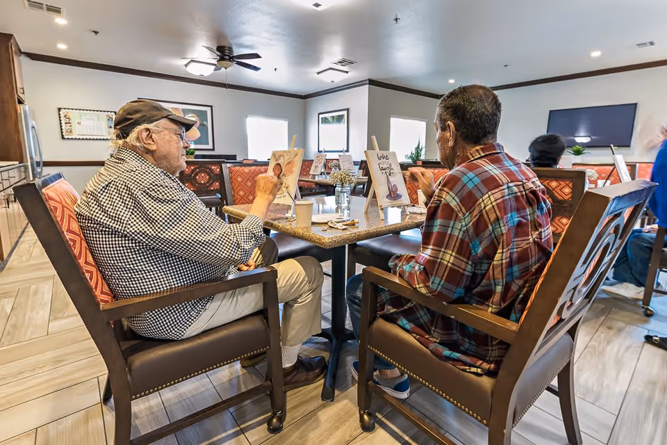 Two elderly men sitting at a table in a well-lit room, engaging in a painting activity with small easels and art supplies. The room has comfortable chairs with patterned cushions, ceiling fans, and windows letting in natural light.