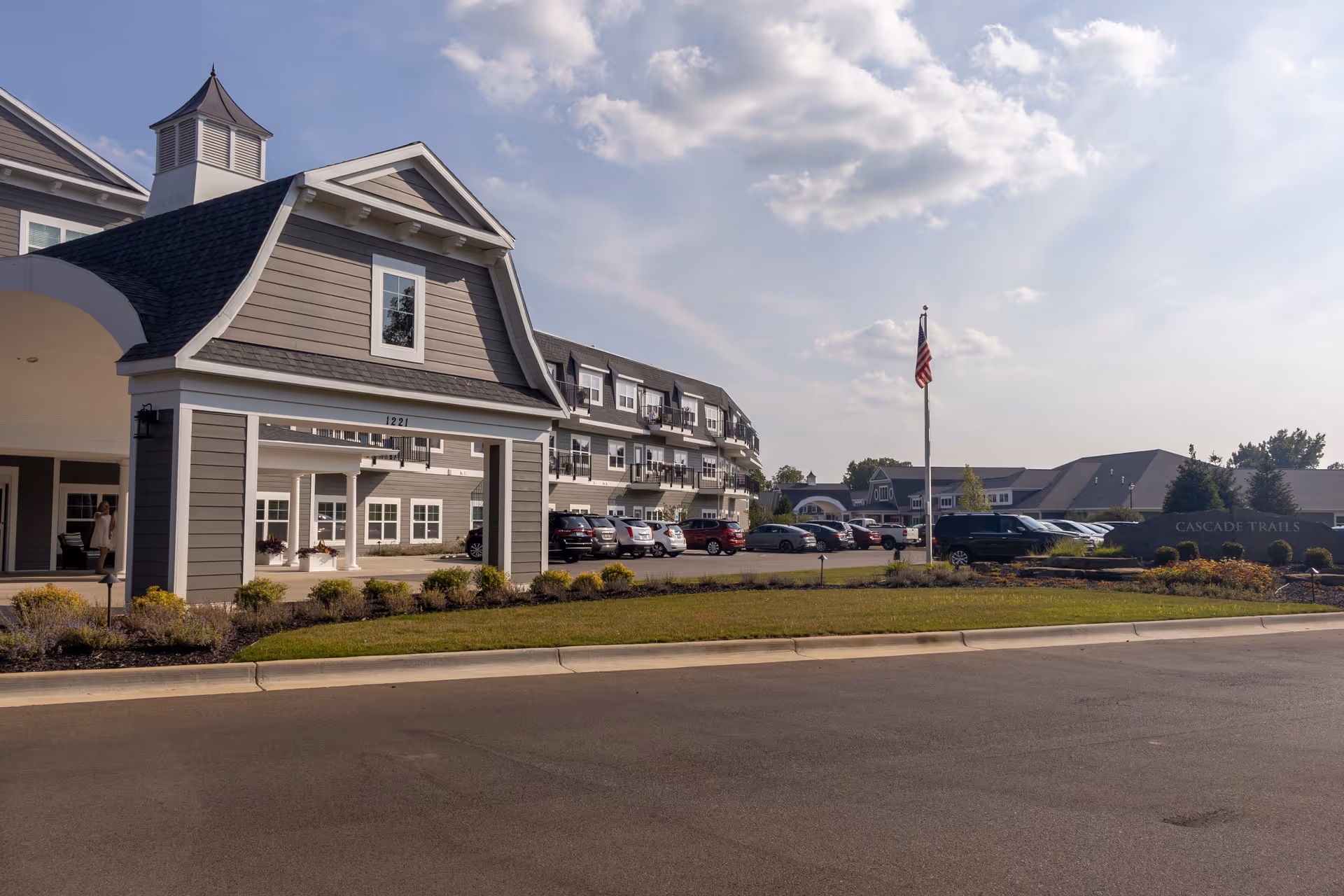 Exterior view of Cascade Trails Senior Living facility showing a large building with multiple windows and balconies, a covered entrance with the number 1221, a parking lot with several cars, an American flag on a flagpole, and a landscaped area with a sign that reads 'Cascade Trails'. The sky is partly cloudy with sunlight illuminating the scene.
