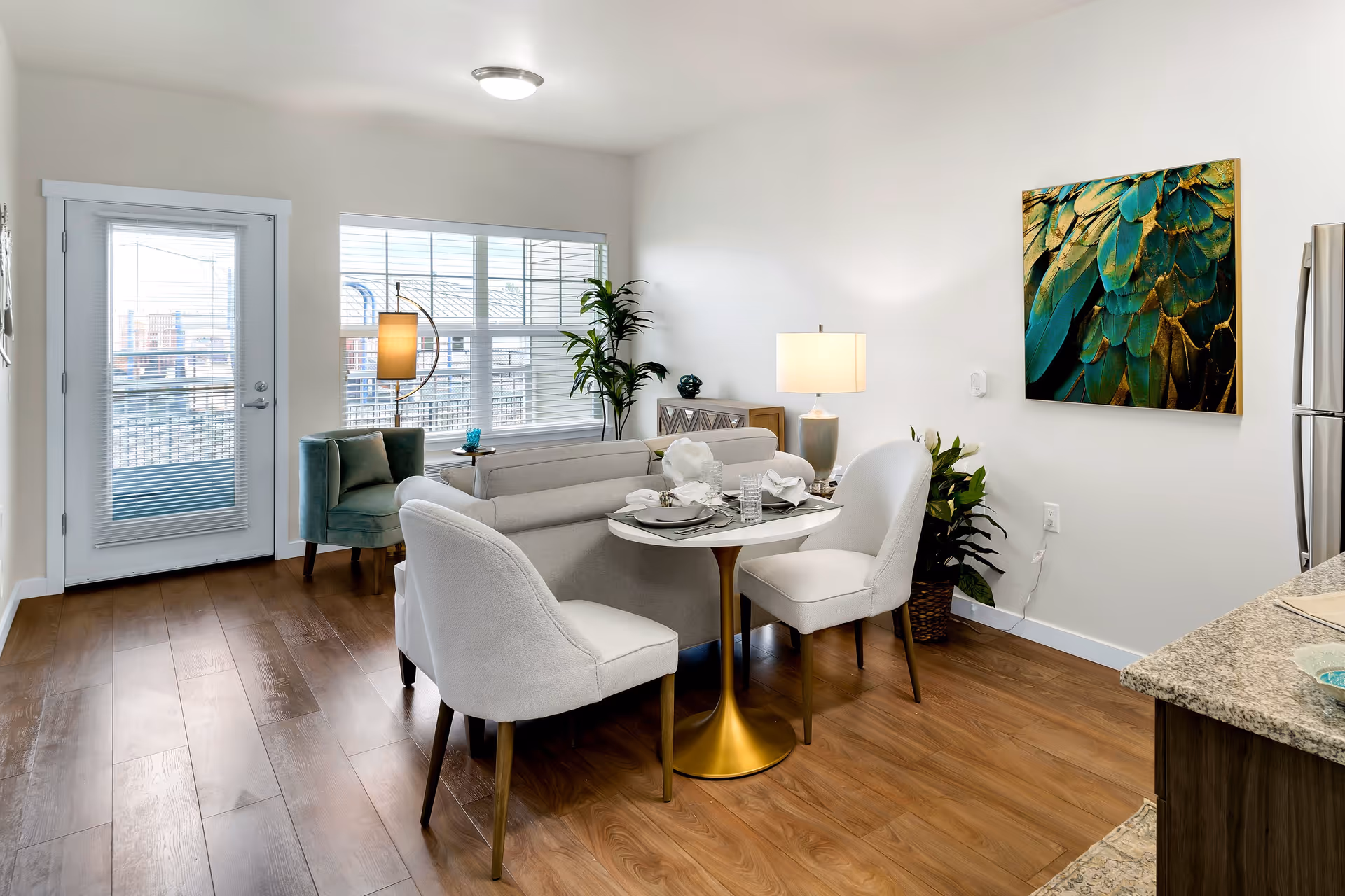 A bright and modern living and dining area with wooden flooring. The space includes a small round dining table with two white upholstered chairs, a light gray sofa, a teal armchair near a window with blinds, a floor lamp, a table lamp on a side table, and a large colorful artwork featuring green and gold feathers on the wall. A glass door leads outside, and part of a kitchen counter with a granite top and stainless steel refrigerator is visible on the right.