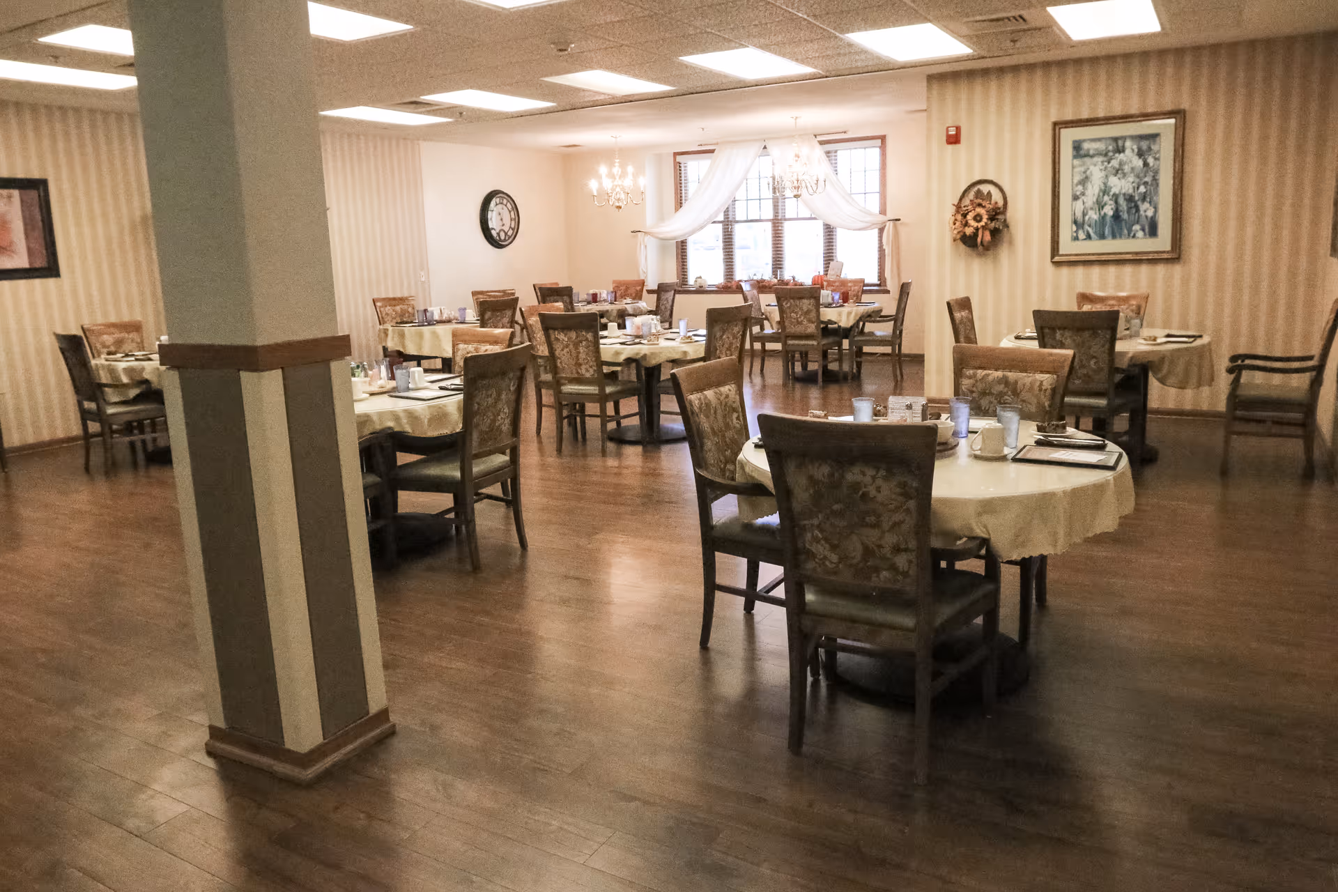 A dining room with several round tables covered with beige tablecloths, each set with cups, glasses, and napkins. The room has wooden flooring, beige striped wallpaper, framed artwork on the walls, and a window with sheer white curtains. There are upholstered chairs around the tables and chandeliers hanging from the ceiling.