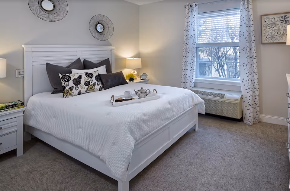 Bright, neatly made white bed in a furnished bedroom with bedside tables, lamps, decorative pillows, and a window with patterned curtains.