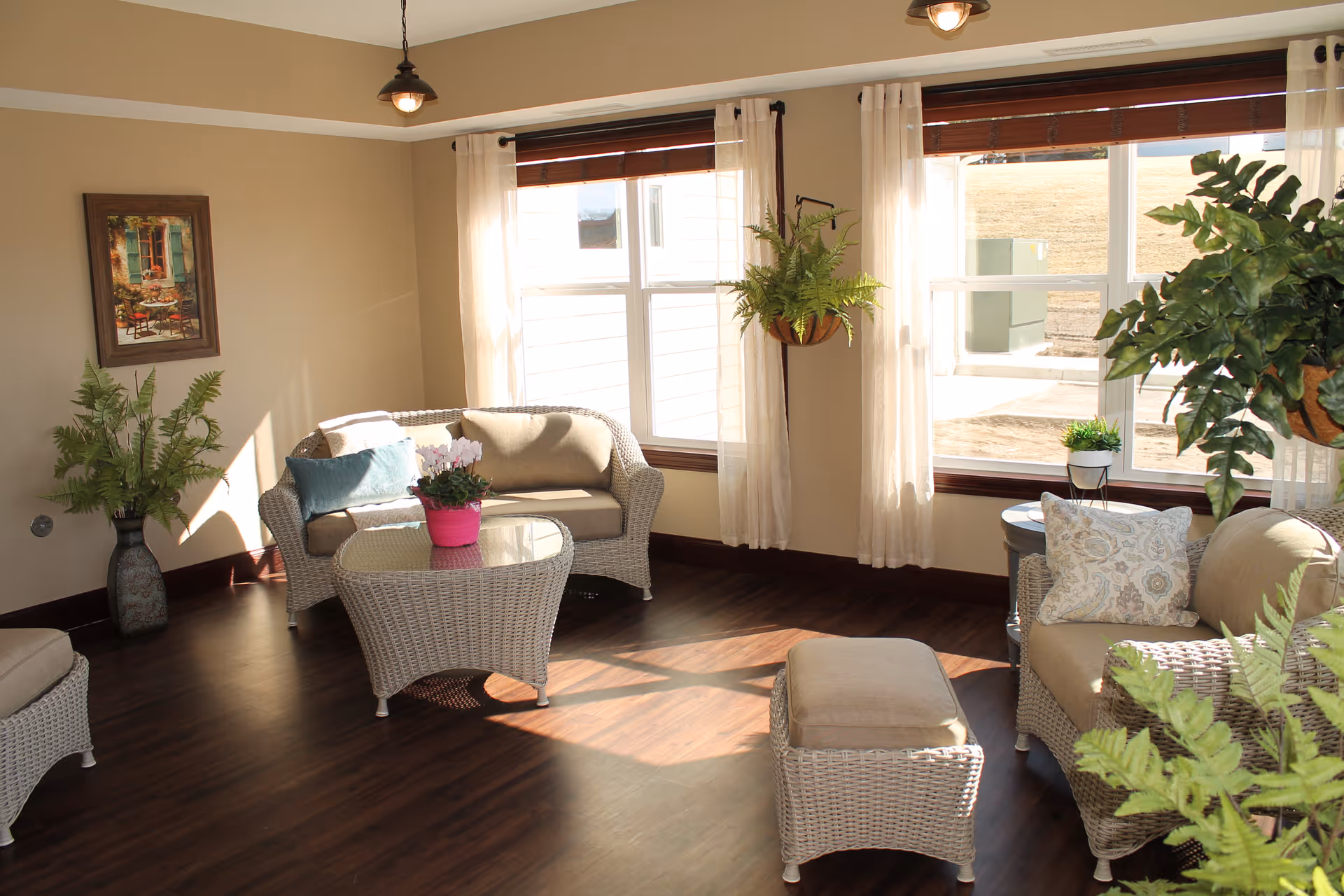 A bright and cozy living room with wicker furniture including a loveseat, armchair, ottoman, and glass-top coffee table. The room has large windows with sheer white curtains allowing natural light to fill the space. There are several green plants placed around the room and a framed painting on the beige wall.