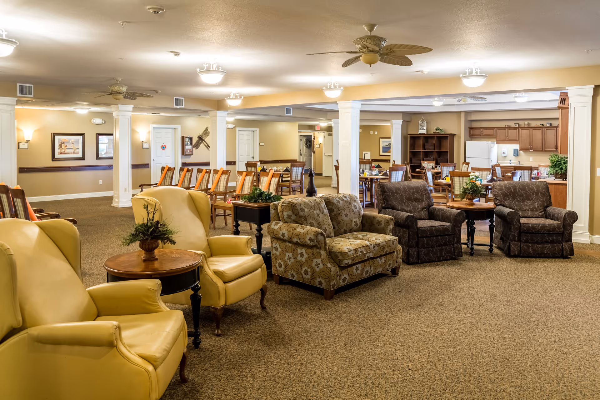 A spacious senior living common area with several comfortable armchairs and sofas arranged around small tables. In the background, there are dining tables and chairs, a kitchen area with cabinets, a refrigerator, and a microwave. The room is well-lit with ceiling lights and ceiling fans, and decorated with plants and framed pictures on the walls.