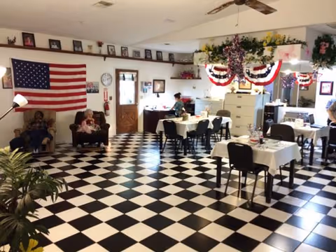 A decorated senior living common/dining room with a black-and-white checkered floor, tables and chairs, an American flag, and residents seated near the wall.