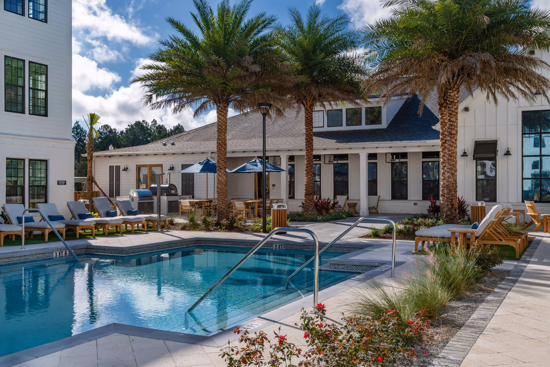 Outdoor pool area with lounge chairs, palm trees and a clubhouse building under blue sky.