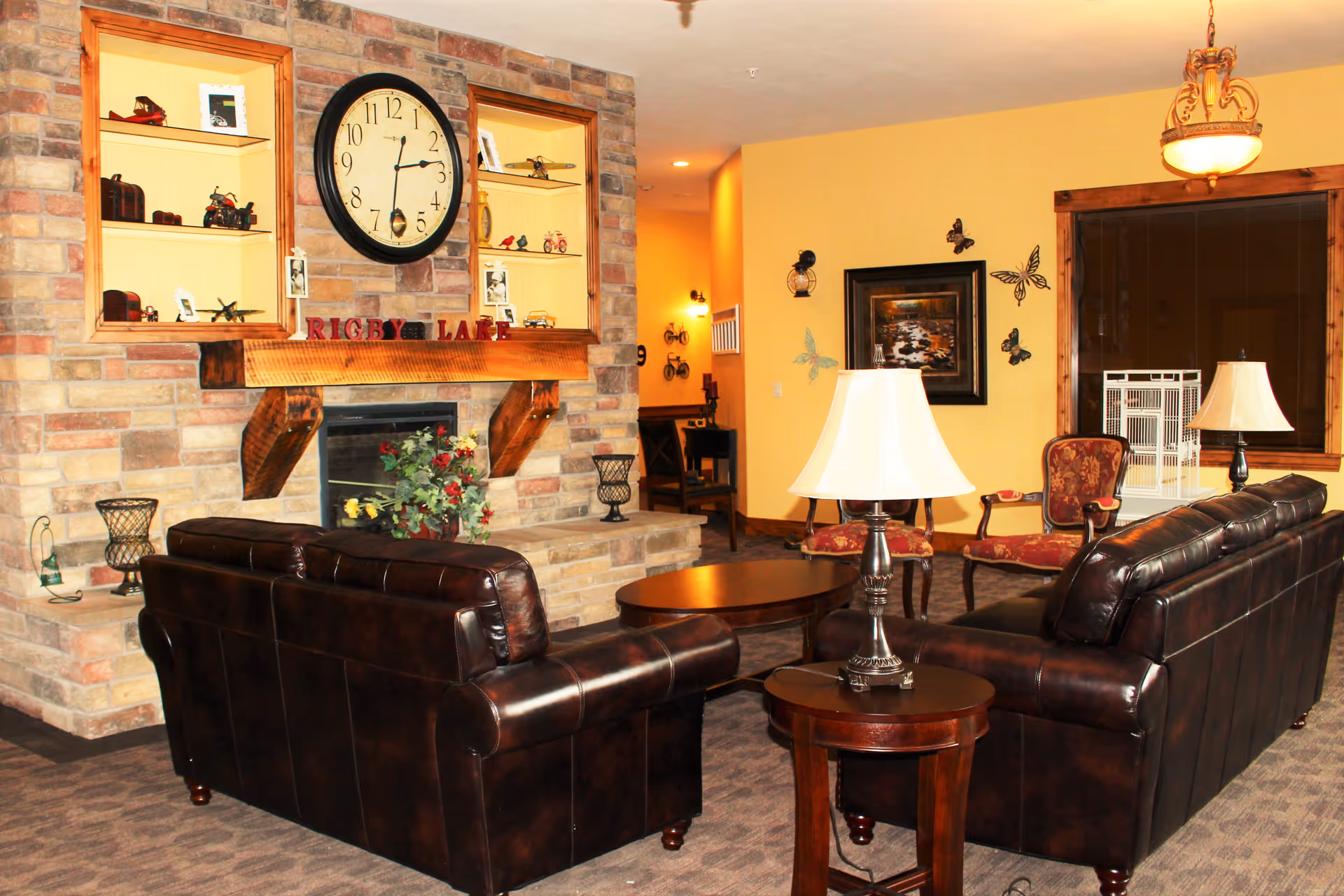 Cozy common living room with dark leather sofas arranged around a stone fireplace topped by a large clock and decorative shelving.