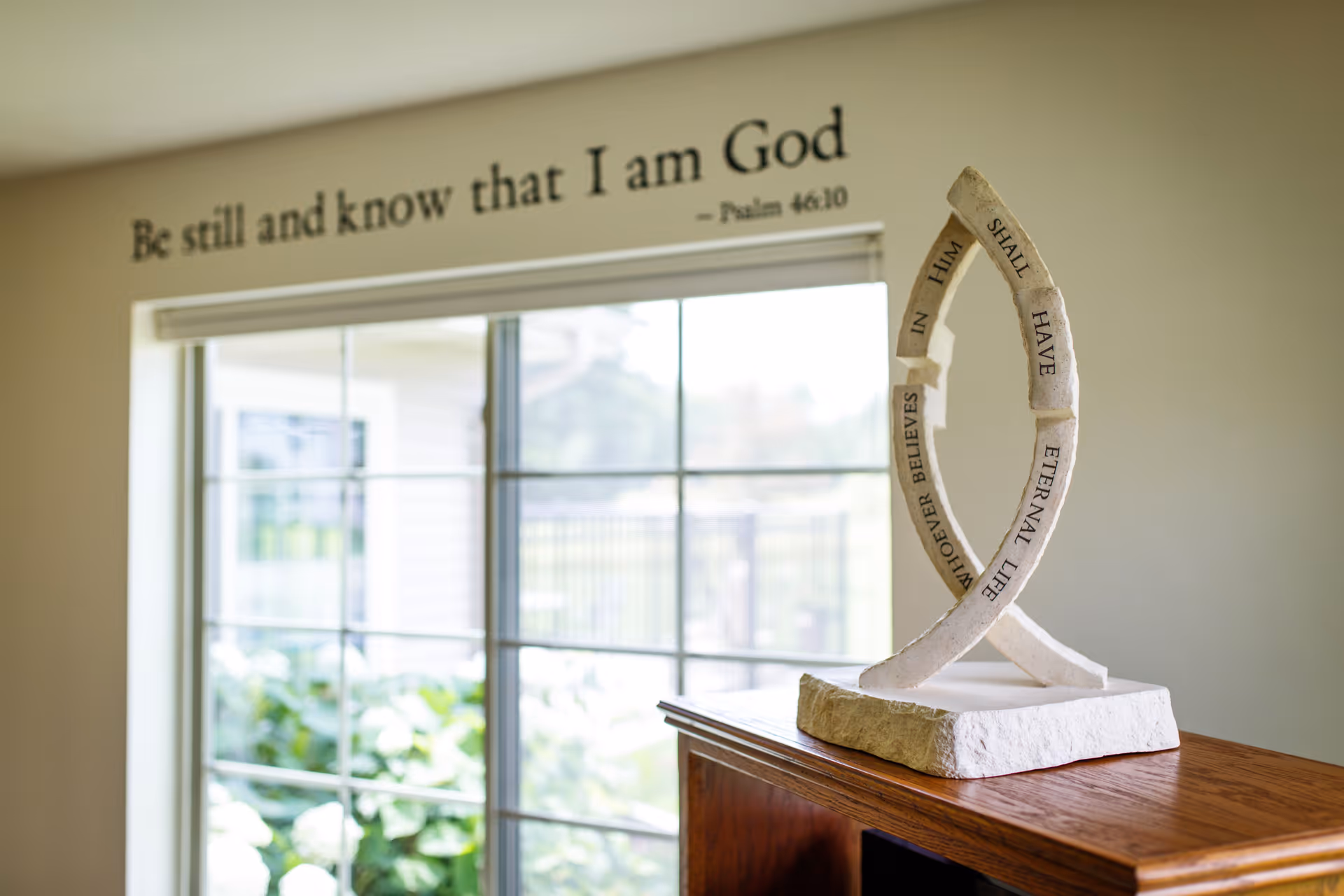 A decorative sculpture with an inspirational religious message sits on a wooden surface near a large window. Above the window, a wall decal reads, 'Be still and know that I am God - Psalm 46:10'. Outside the window, greenery and a fence are visible.