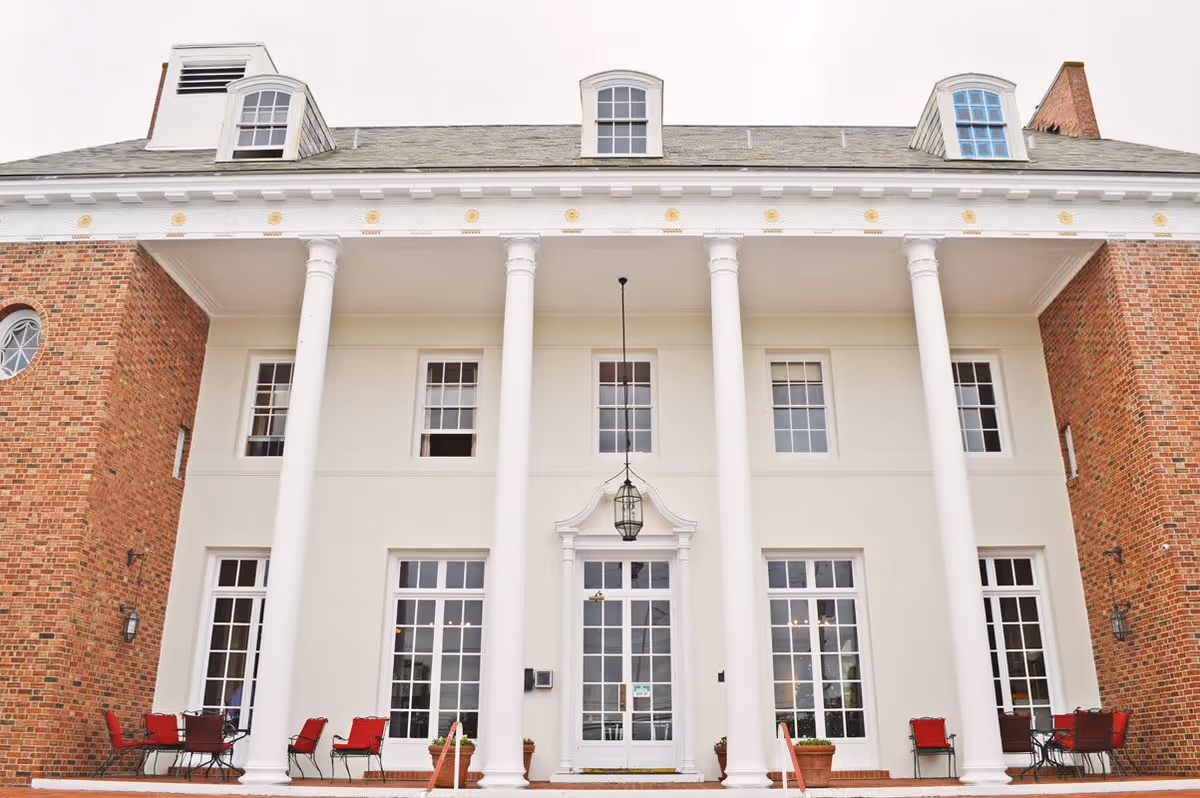 Front exterior view of a large building with white columns and brick walls, featuring multiple windows and a central door with glass panels. There are red cushioned chairs and tables arranged on the porch area.