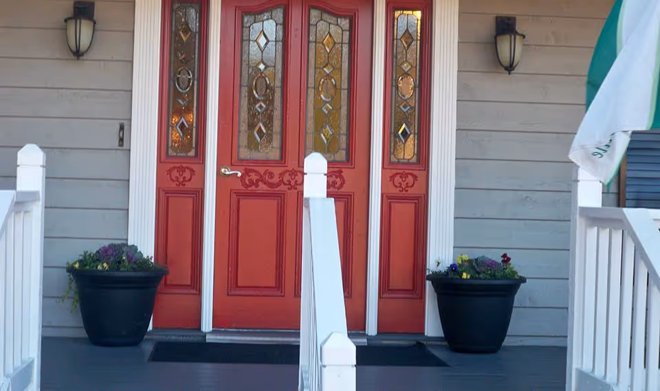 Front entrance of a building with a red door featuring decorative glass panels. There are two black planters with flowers on either side of the door, white railings leading up to the entrance, and two wall-mounted lantern-style lights on either side of the door.