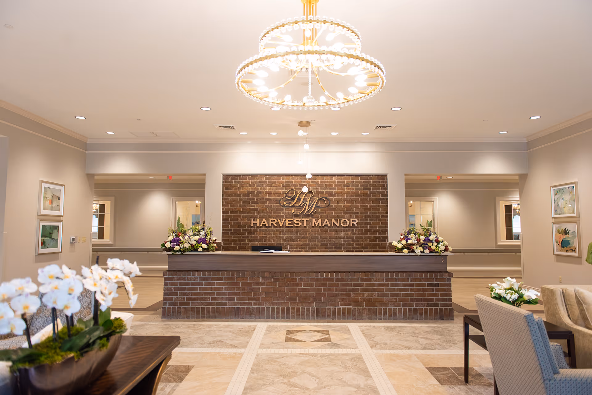 Well-lit reception lobby with a brick front desk reading "Harvest Manor", a chandelier overhead, and floral arrangements.