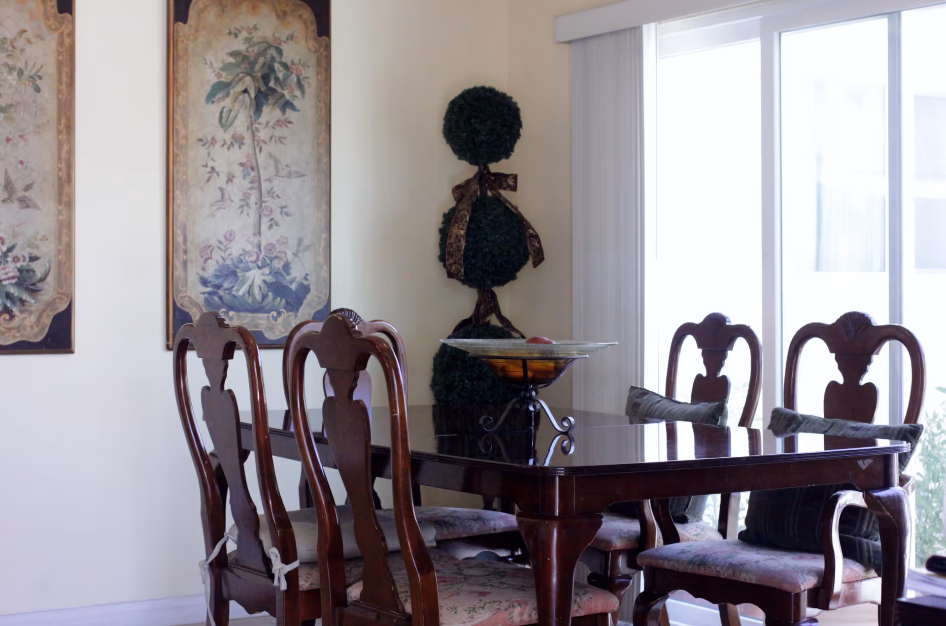 A dining room with a dark wooden table and six matching chairs with floral cushions. On the table is a decorative bowl with fruit. Behind the table are two framed floral paintings on the wall and a topiary plant with a ribbon. Large sliding glass doors with vertical blinds let in natural light.