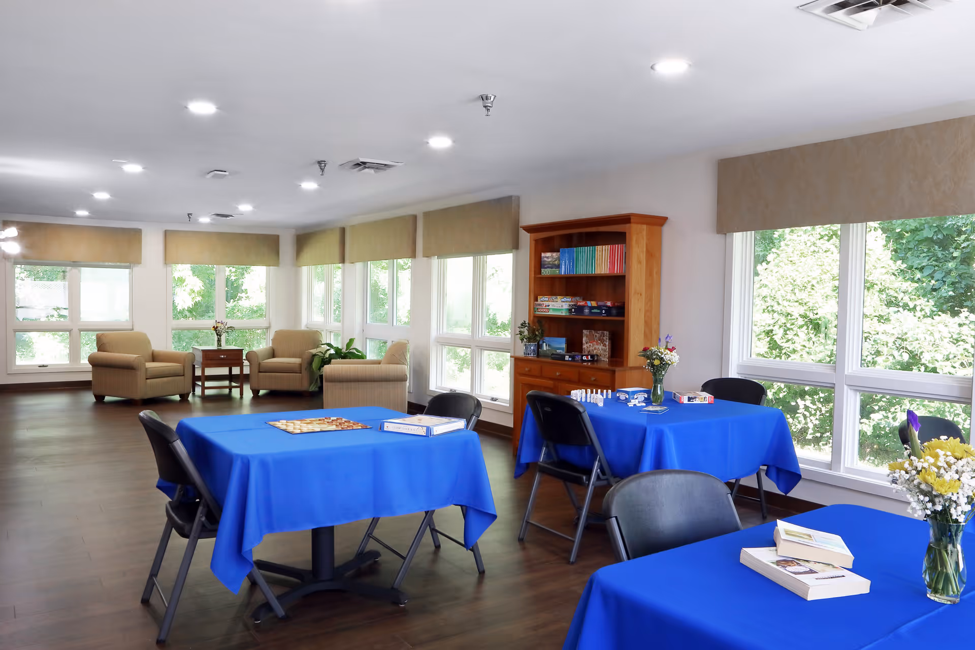 A bright and spacious common area with large windows letting in natural light. The room has several tables covered with blue tablecloths, each with chairs around them. On the tables are board games, books, and flower vases. In the background, there are comfortable armchairs and a wooden bookshelf filled with board games and books. The floor is wooden, and the walls are white with beige window valances.