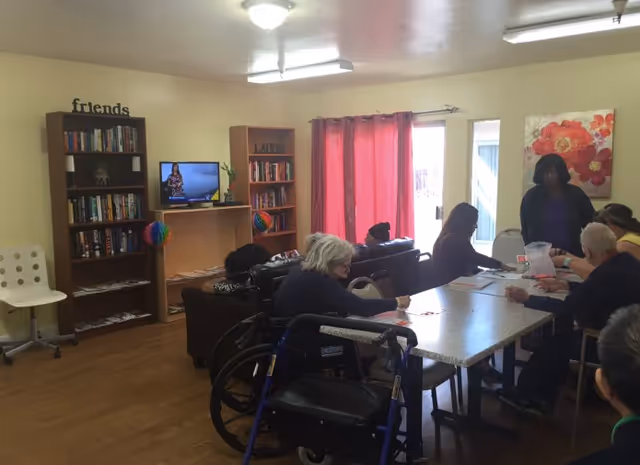 A common area in a senior living facility with several elderly people sitting around a table engaged in activities. There are bookshelves filled with books, a television, and a couch in the background. The room has wooden flooring, light yellow walls, and red curtains covering a window or door. A walker is visible near one of the seated individuals.