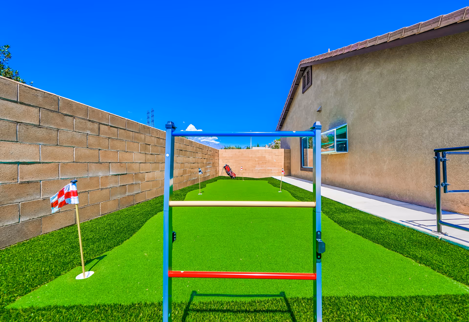 Outdoor putting green area with artificial grass, surrounded by a brick wall and the side of a building. There are small golf flags and a golf bag in the background, along with a ladder toss game frame in the foreground.