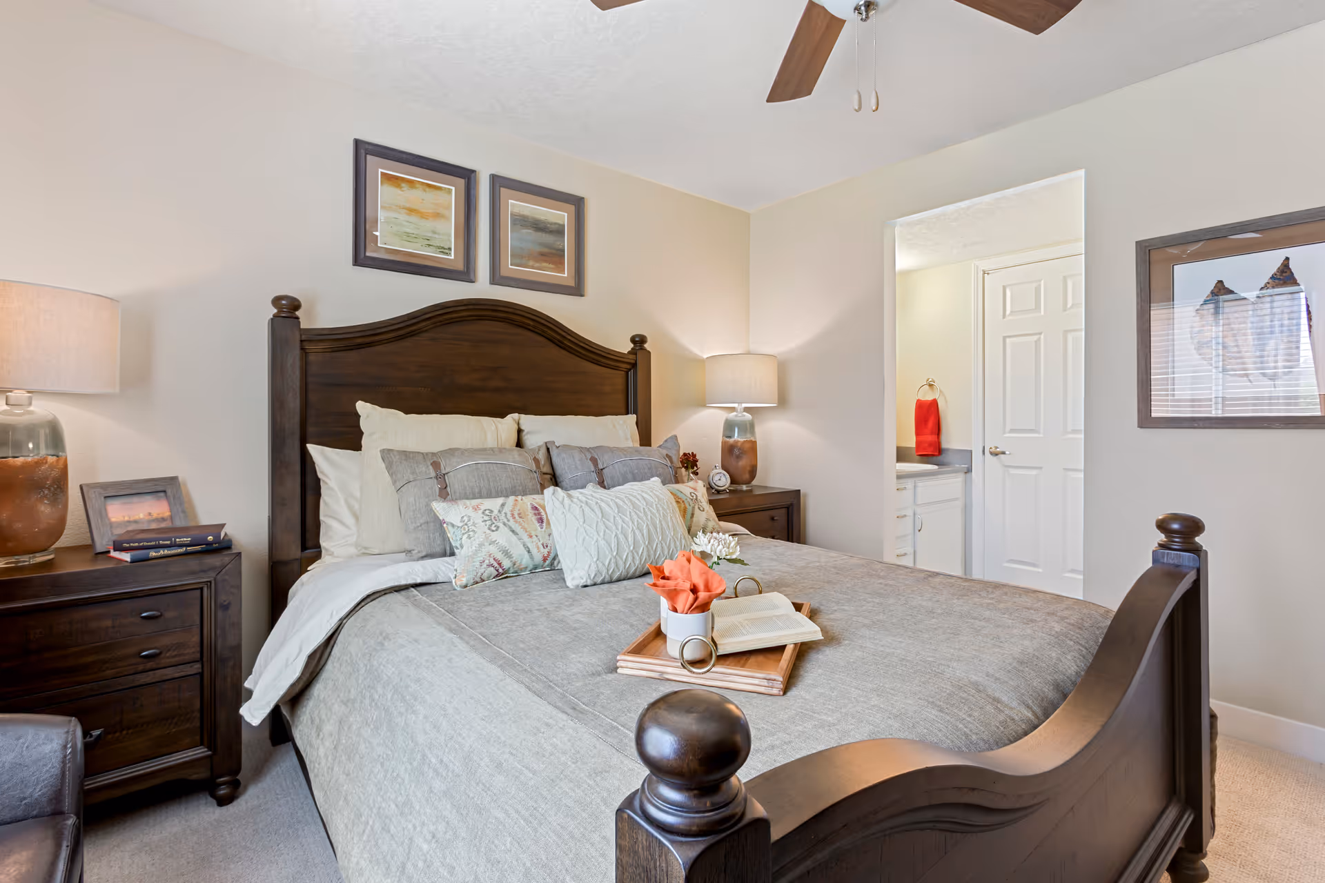 A cozy bedroom with a dark wooden bed frame and headboard, neatly made bed with multiple pillows and a gray bedspread. There are two nightstands on either side of the bed, each with a lamp. A tray with a book, a cup, and a flower is placed on the bed. The room has light-colored walls with two framed pictures above the headboard and a framed picture on the right wall. An open doorway leads to a bathroom with a white door and a red towel hanging inside.