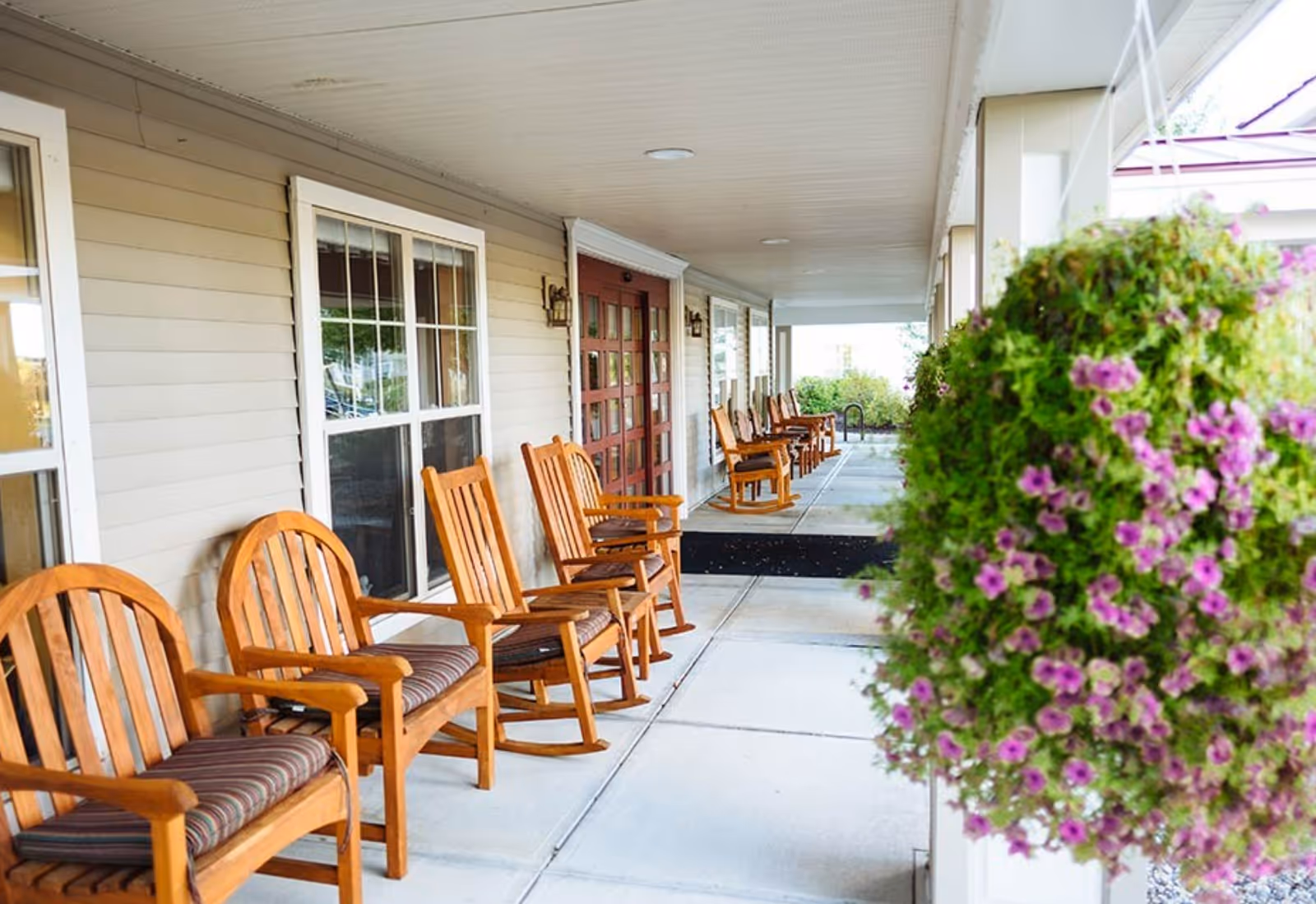 Covered porch area with a row of wooden rocking chairs with cushions lined up against the wall of a building. There are windows and a double door along the wall, and a hanging basket with green foliage and purple flowers is visible in the foreground on the right side.